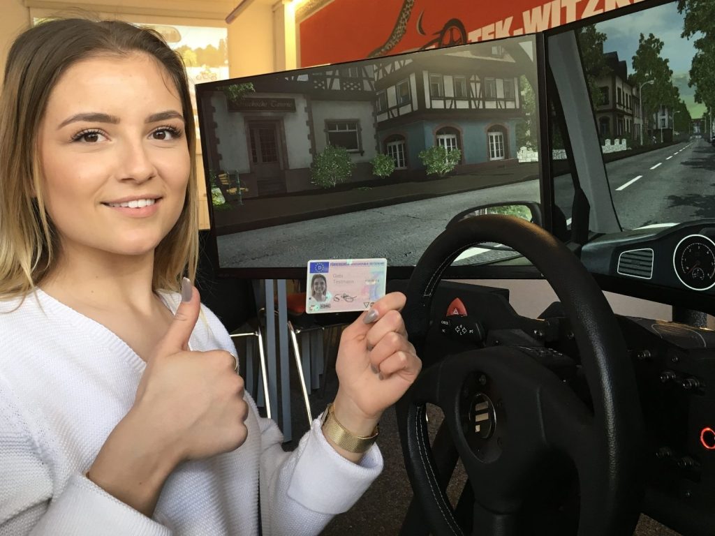 Ein junges Mädchen sitzt im Fahrsimulator und har ihren Führerschein bestanden. Sie hält den Führerschein nach oben.